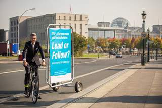 ProTTIP Plakatoffensive in Berlin BDI-Präsident Ulrich Grillo im Berliner Regierungsviertel auf einem Fahrrad mit Anhänger, auf dem ein Plakat der ProTTIP-Offensive mit dem Slogan „Lead or Follow?“ angebracht ist.