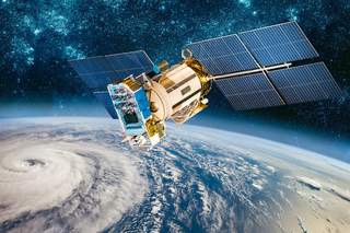 Ein Satellit mit zwei aus Solaranlagen bestehenden Flügeln, kreist um unsere Erde. Die Erde ist als blauer Planet mit einer großen weißen Wolkendecke dargestellt. Der Weltraum im Hintergrund ist dunkelblau und es sind weiß funkelnde Sterne zu sehen.